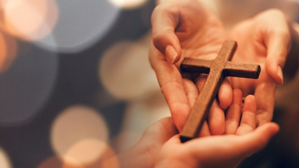Das Bild zeigt ein kleines Kreuz aus Holz, das von drei Händen gehalten wird.